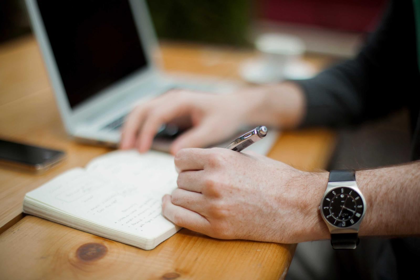 Hands writing notes in a notebook beside a laptop